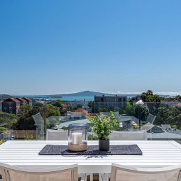 A 6 seater white outdoor dining table remains low line to showcase the view over Rangitoto Island.
